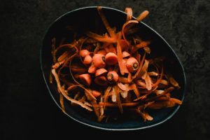 An image of peeled carrots, and cut off carrot tops, in a bowl on a dark background representing food waste.