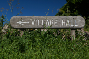 Image shows a wooden signpost for a village hall, standing in a green hedge against a blue sky background.