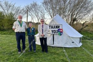 A photo of a Scout Leader, Scout, and the Chair of South Brent Parish Council, standing in front of a white bell tent in a field. A thank you sign is hung on the tent.